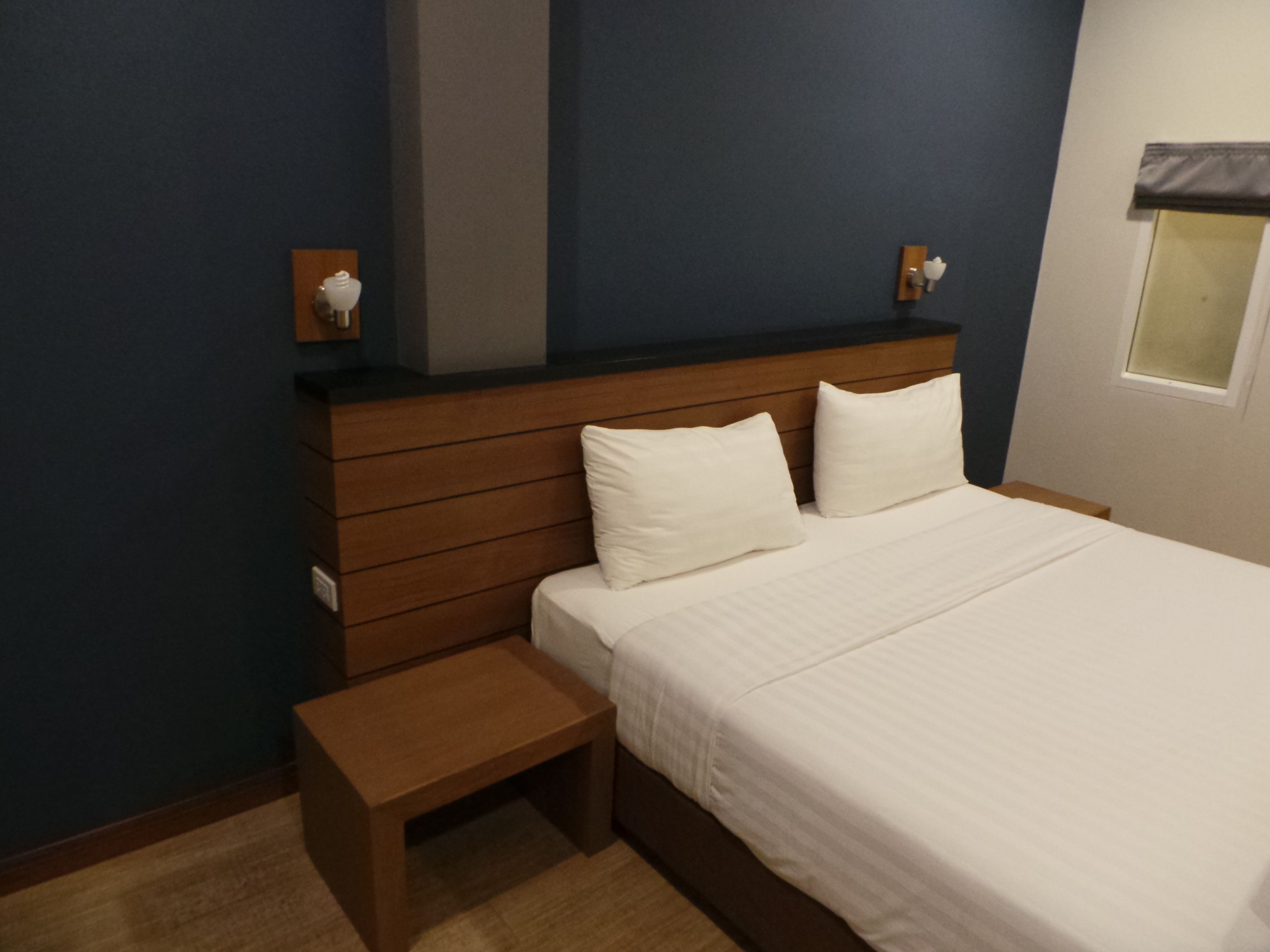 A bedroom with a wooden headboard, a single bed with white bedding, a small wooden bench, and a nightstand with a lamp.