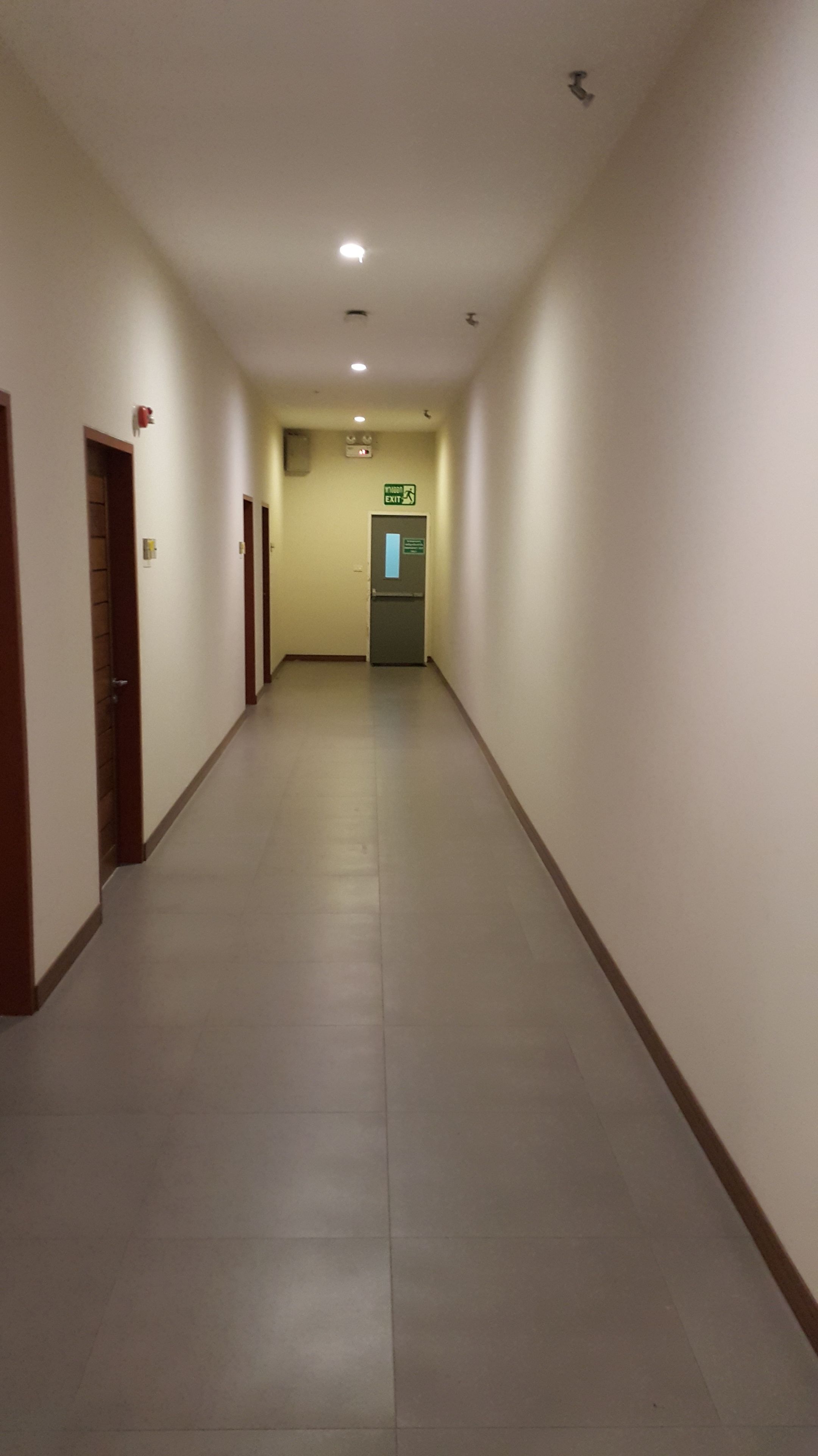 Um corredor comprido e estreito com chão de azulejos e paredes brancas. Há portas em ambos os lados e uma placa de saída verde é visível ao longe.
