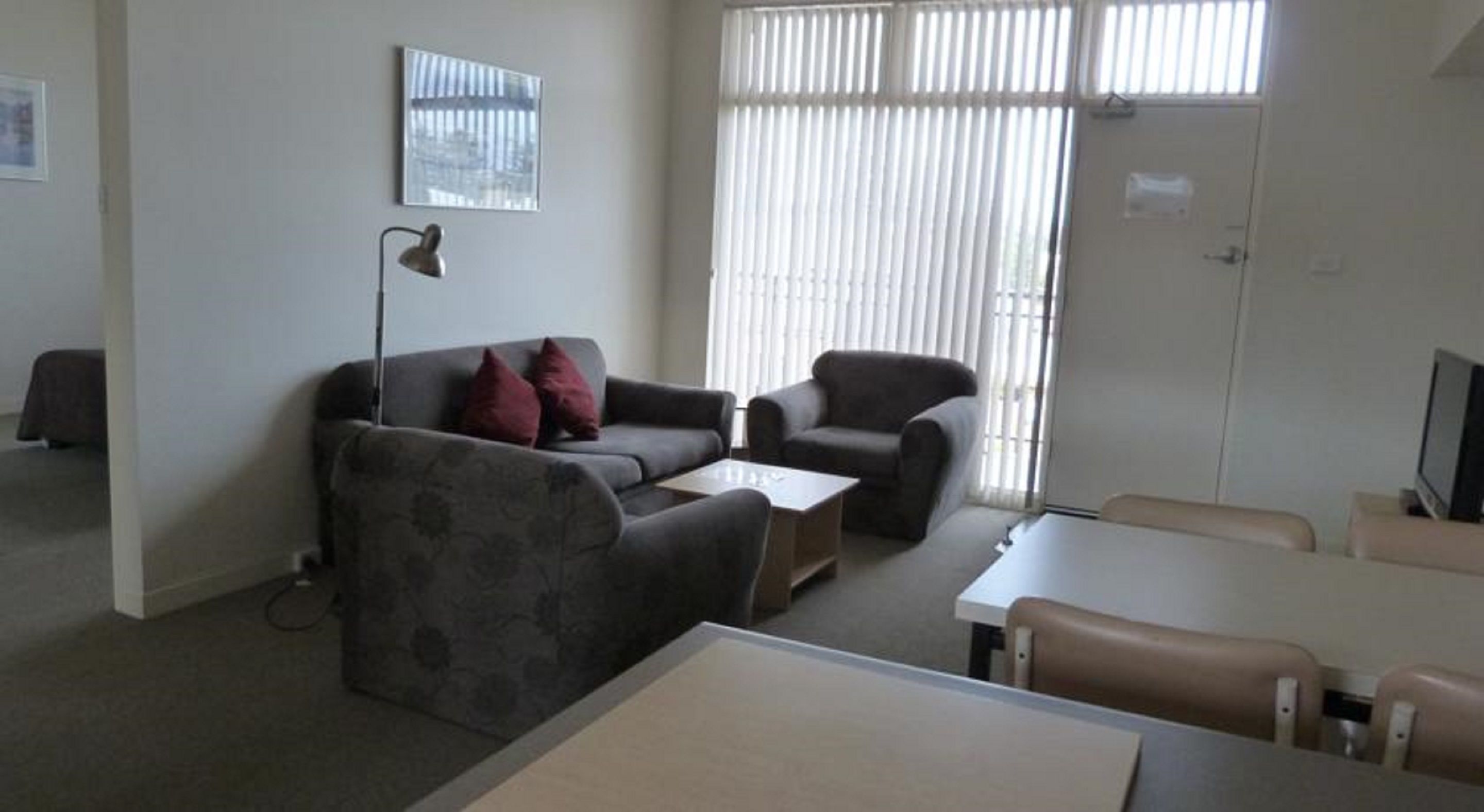 A living room with a sofa, armchair, and a small table. There is a lamp, a TV, and a window with blinds.