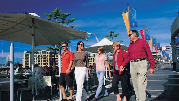 A group of people walking on a waterfront promenade with outdoor seating and umbrellas.