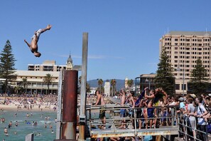 Una plataforma pública de buceo con gente alrededor, una playa y un edificio alto al fondo.