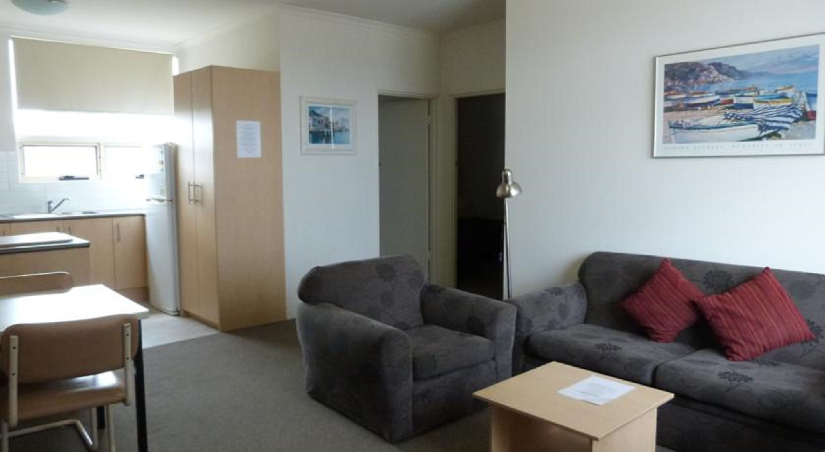 Un espacio habitable compacto con kitchenette, dos sillones, un sofá con cojines rojos y un cuadro enmarcado en la pared.