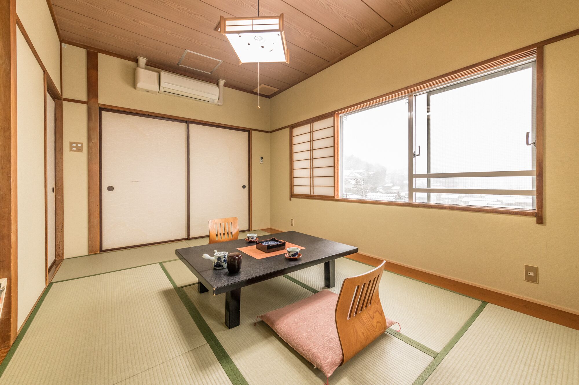 Una habitación japonesa tradicional con suelo de tatami, una mesa baja, una silla acolchada, puertas correderas y un ventanal con vistas.