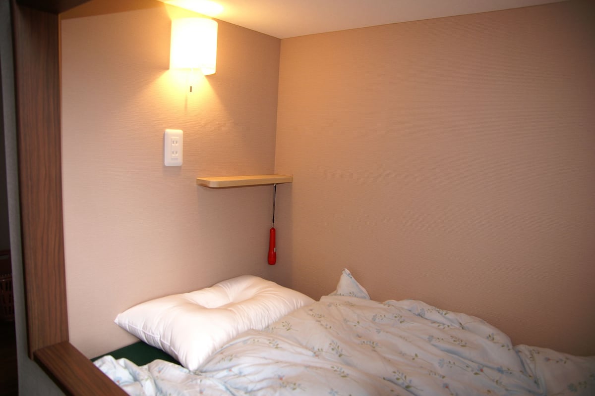 A small, neatly made bed with a white pillow and floral-patterned bedding, a bedside table with a red object, and a wall-mounted light fixture.