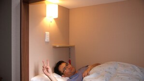 A person lying in a bed with a white pillow and a floral-patterned blanket. There is a wall-mounted light fixture above the bed.