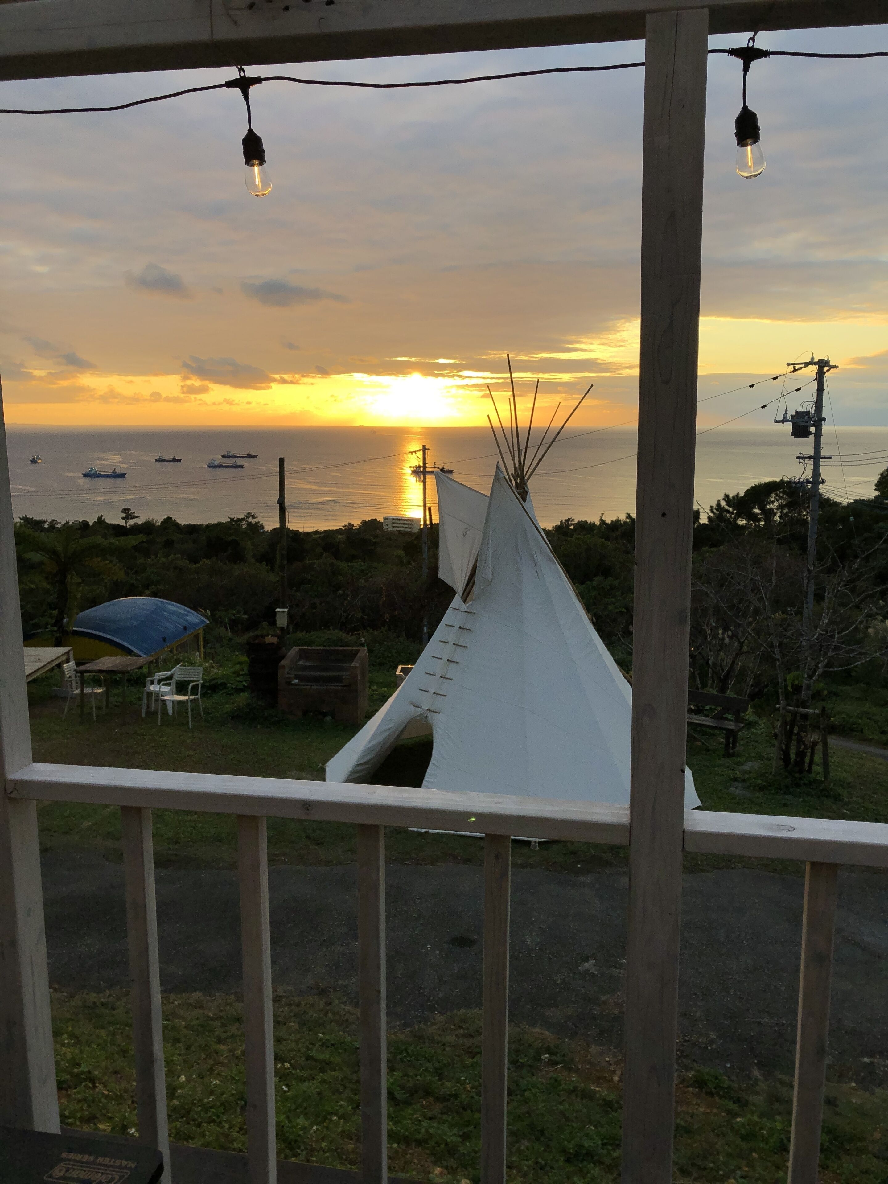 Vista del atardecer sobre el océano desde un porche con barandilla de madera, una tienda tipi blanca y un paraguas azul.