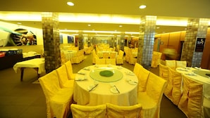 Un salón de banquetes con mesas redondas y sillas amarillas, un área de recepción y una pared de azulejos con un mural.