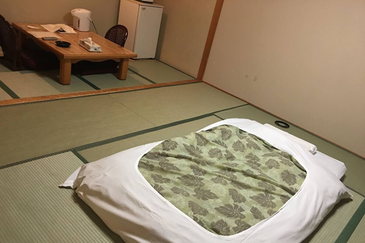 A tatami mat room with a bed, a small table with a kettle, and a refrigerator.