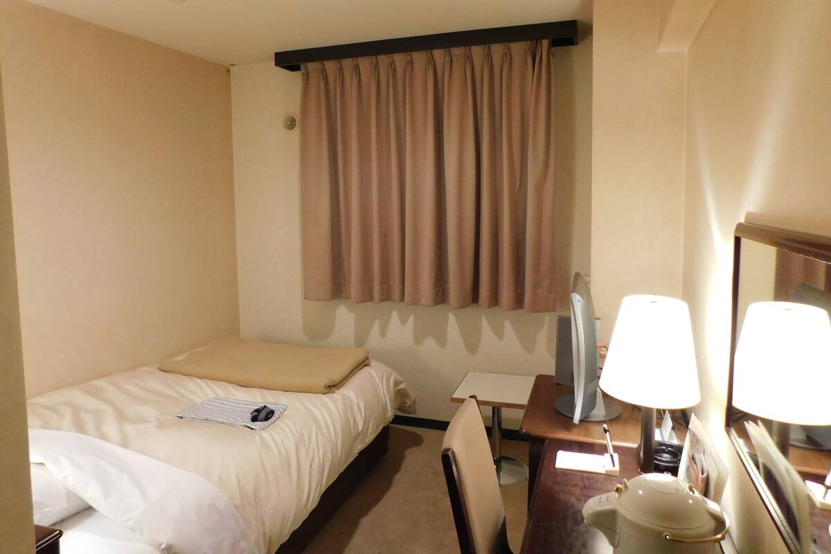 A hotel room with a bed, a desk, a chair, a lamp, and a window with curtains.