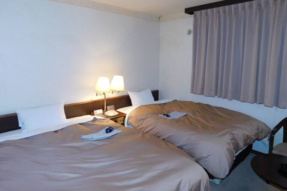 A hotel room with two beds, a nightstand with a lamp, a chair, and a window with curtains.