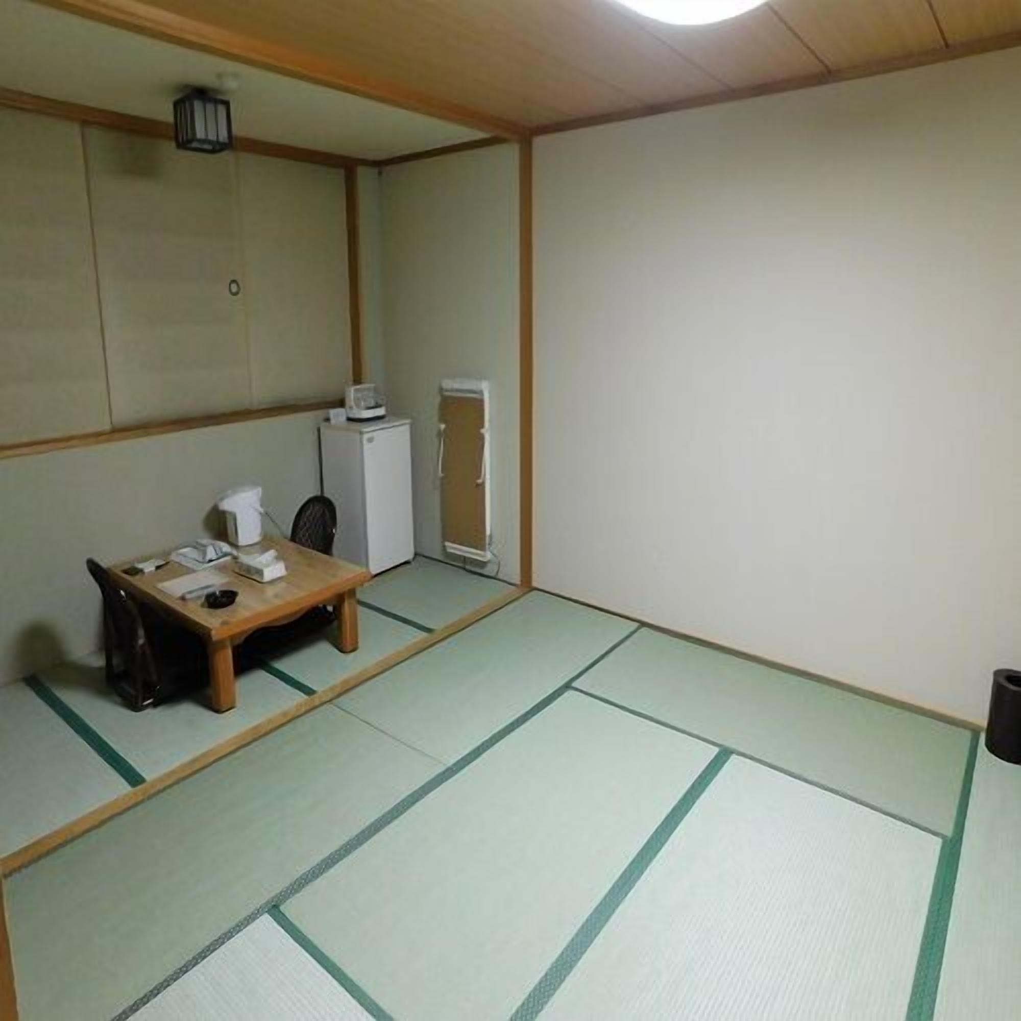 Une pièce de style japonais traditionnel, avec un sol recouvert de tatamis, une petite table basse accompagnée d’une chaise, un petit radiateur et un réfrigérateur.