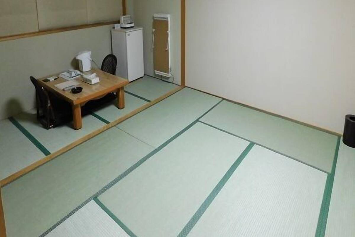 A traditional Japanese room with tatami flooring, a low table with a chair, a small heater, and a refrigerator.