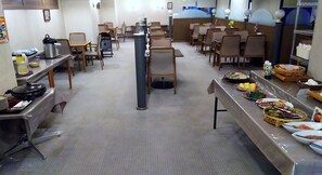 Una cafetería con varias mesas y sillas, equipo de servicio de alimentos y una variedad de platos en una mesa de buffet.