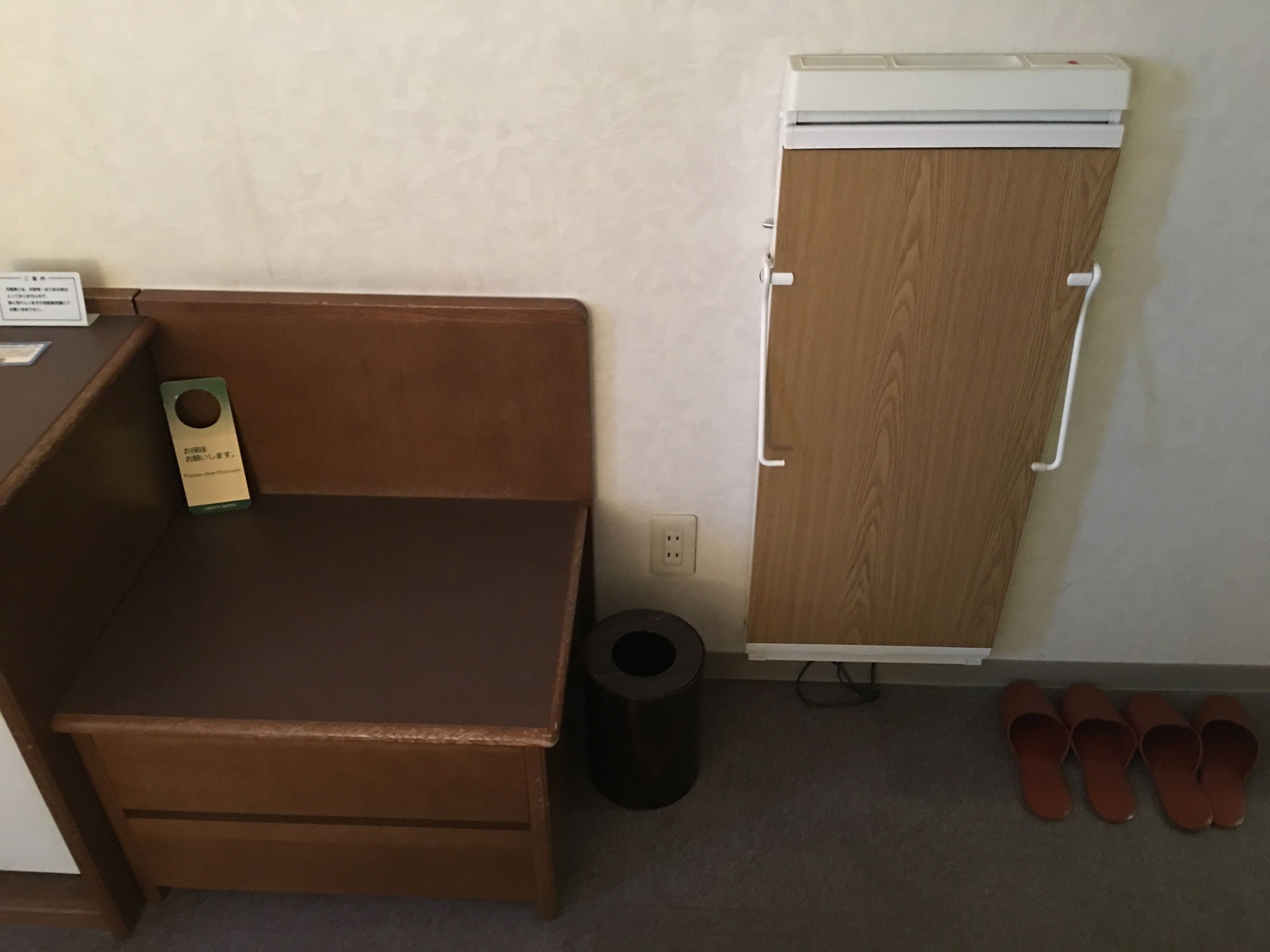 A small room with a wooden desk, a trash can, a white air conditioner, and a pair of slippers on the floor.