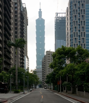A street view with modern buildings, a prominent tower in the background, and trees lining the road.