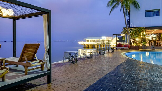 A poolside area with lounge chairs and a covered seating area, a palm tree, and a building with illuminated signage in the background. - The Loft Seaside Sriracha (Si Racha)