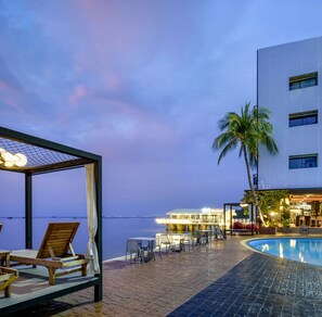 A poolside area with lounge chairs and a covered seating area, a palm tree, and a building with illuminated signage in the background. - The Loft Seaside Sriracha (Si Racha)