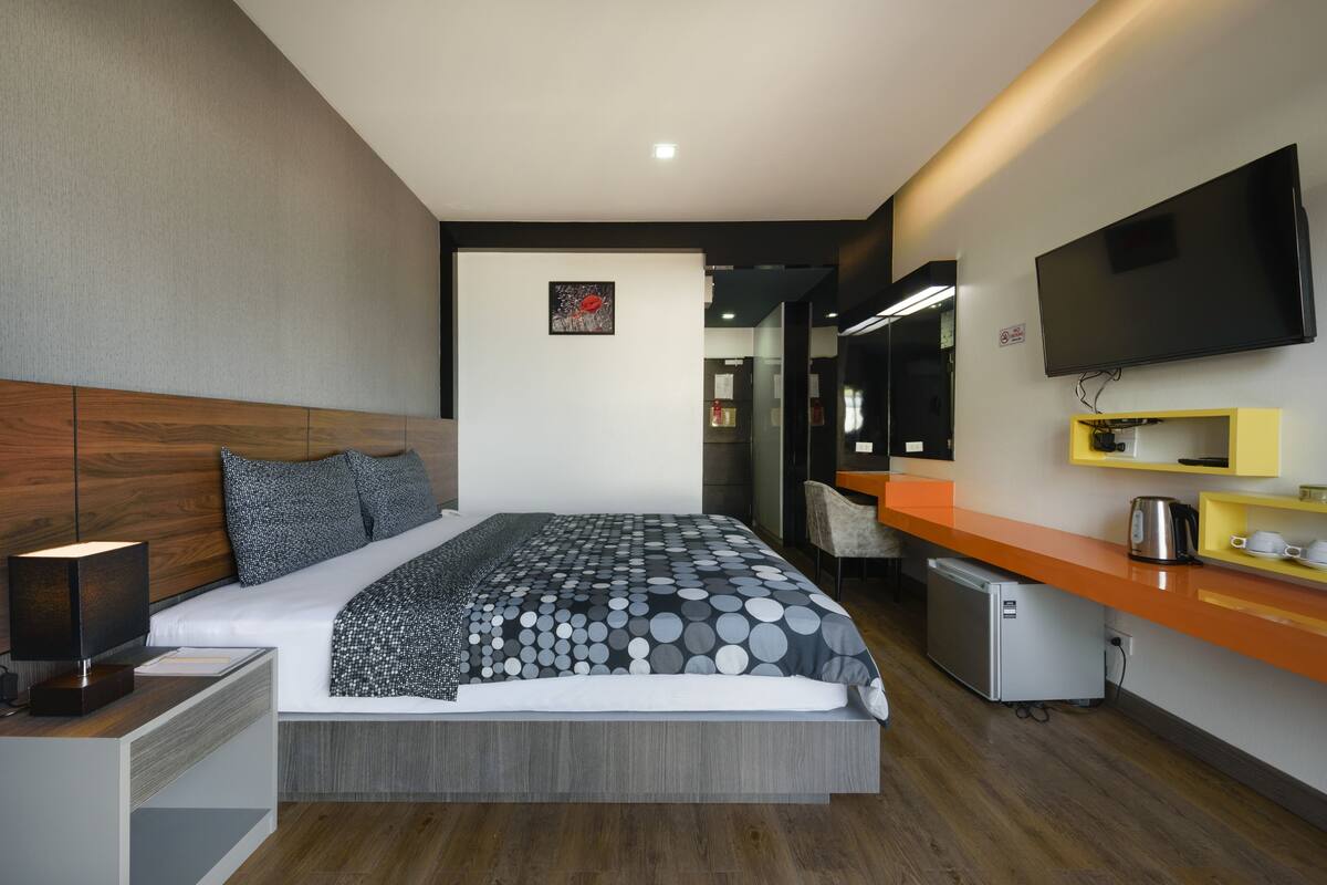 A modern hotel room with a large bed, a flat-screen TV, a small kitchenette, and a bathroom visible through an open door.