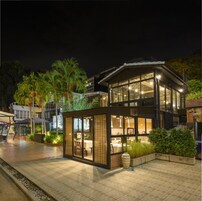 A modern building with large windows and a glass enclosure, surrounded by greenery and a paved area.