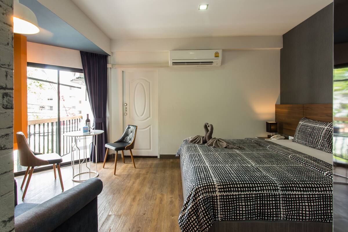 A hotel room with a bed, a chair, a small table, and an air conditioning unit.
