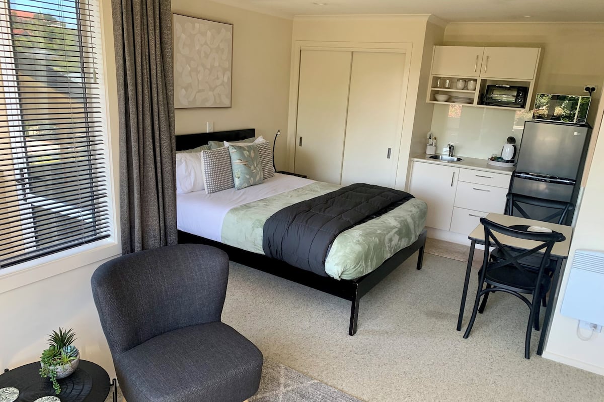 A bedroom with a bed, a chair, a small table with a plant, and a kitchenette with a refrigerator, stove, and microwave.