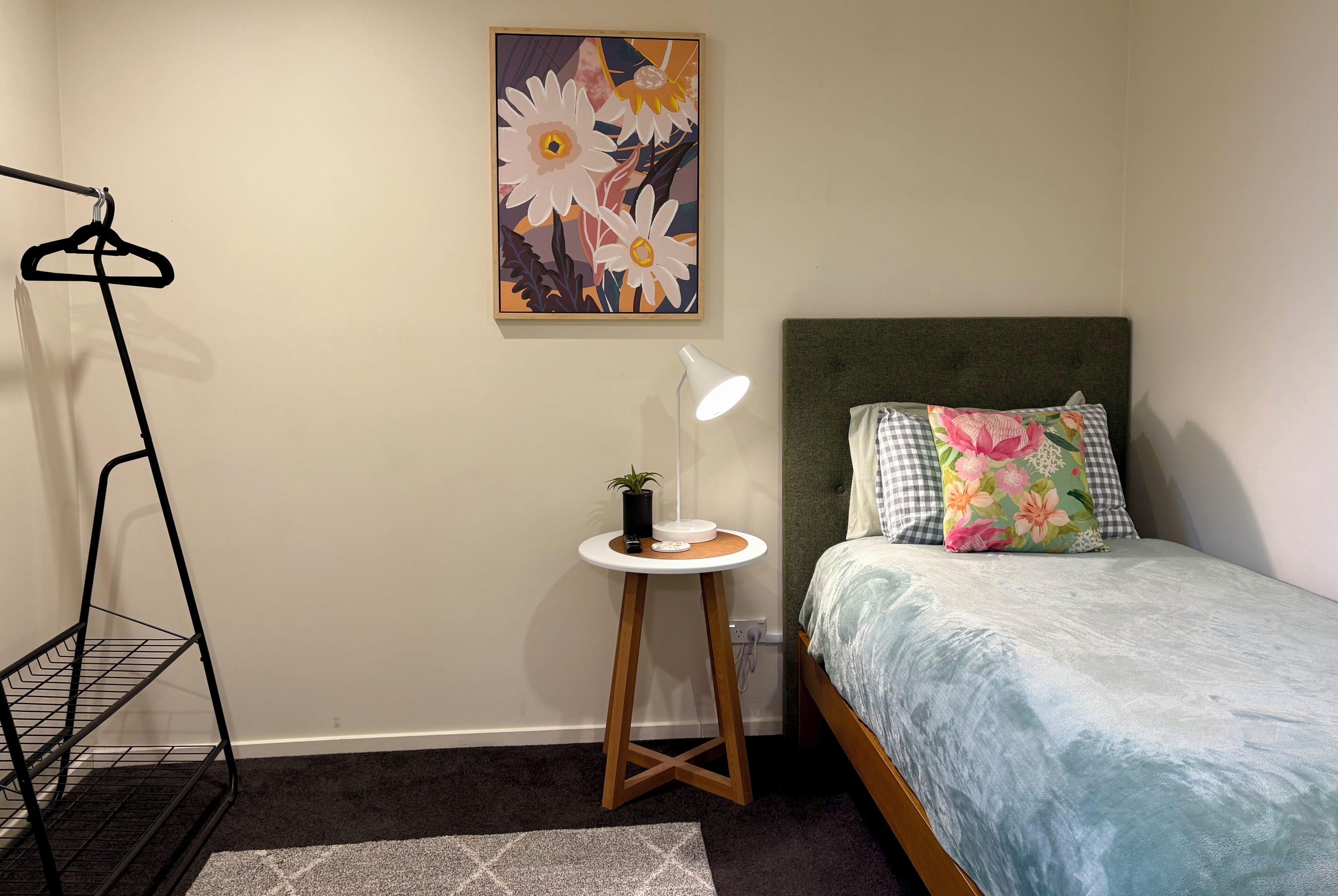 A bedroom with a bed, a bedside table with a lamp, a hanging rack, and a floral painting on the wall.