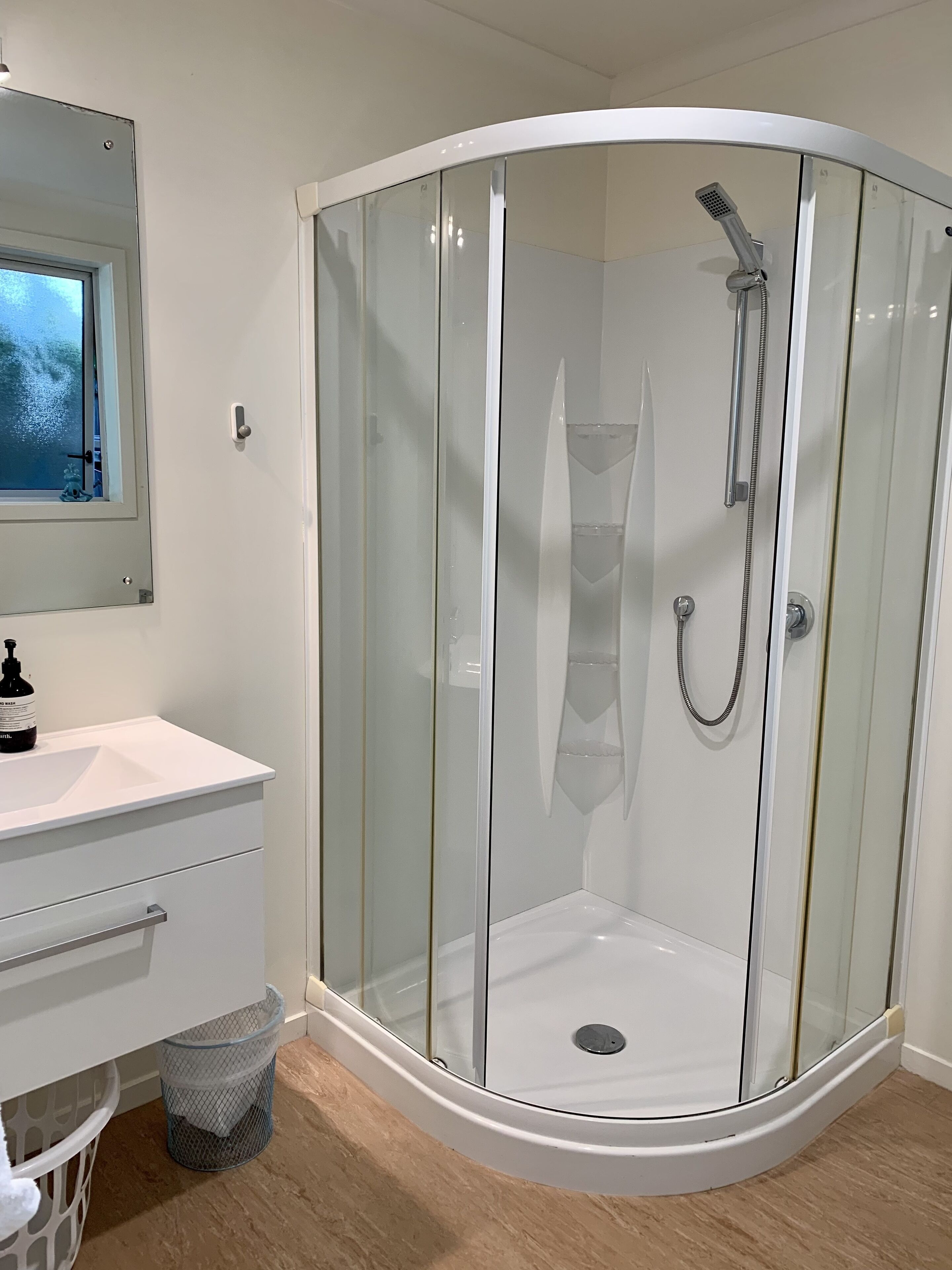 Un baño moderno con ducha envuelta en vidrio, tocador blanco con lavamanos y una ventana sobre el espejo.