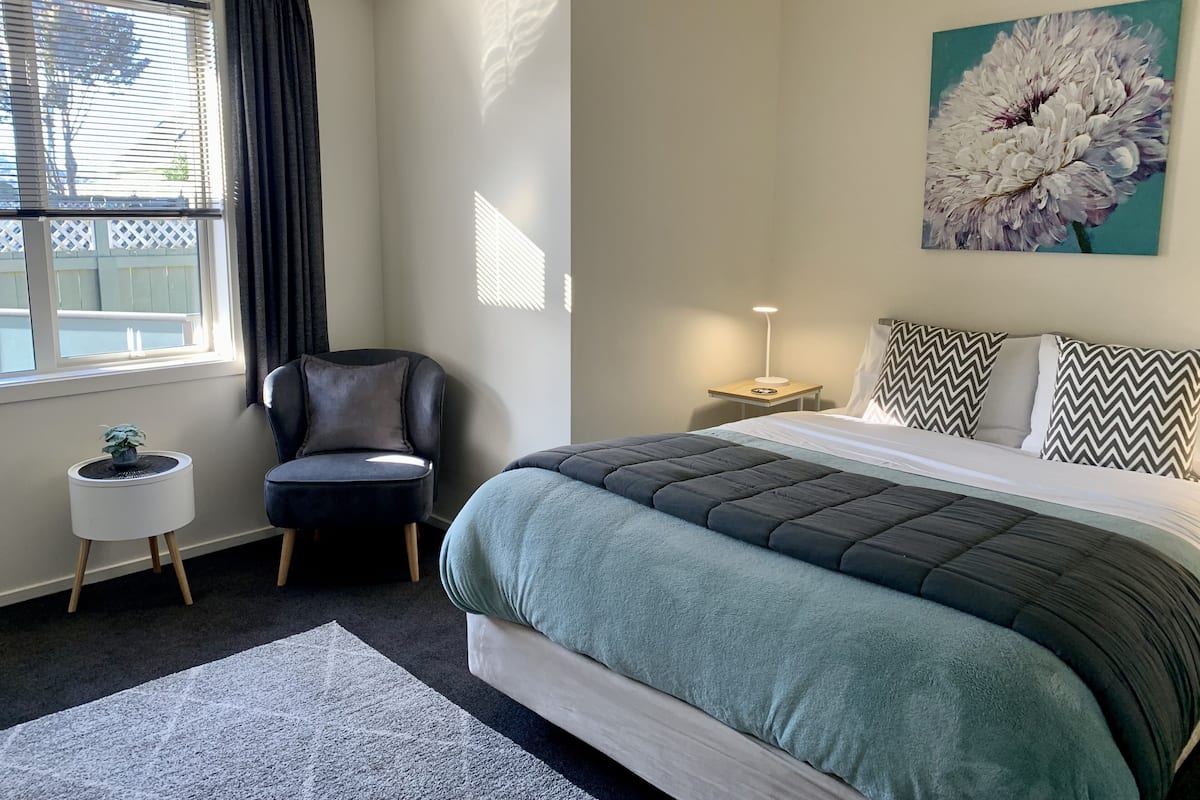 A bedroom with a bed, a chair, a small table, a lamp, and a painting on the wall.