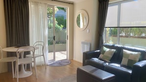 A living room with a black leather sofa, a round coffee table, and a dining table with chairs. There is a sliding glass door leading to a garden.