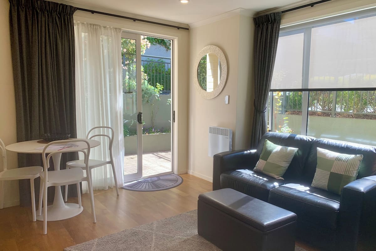 A living room with a black leather sofa, a round coffee table, and a dining table with chairs. There is a sliding glass door leading to a garden.