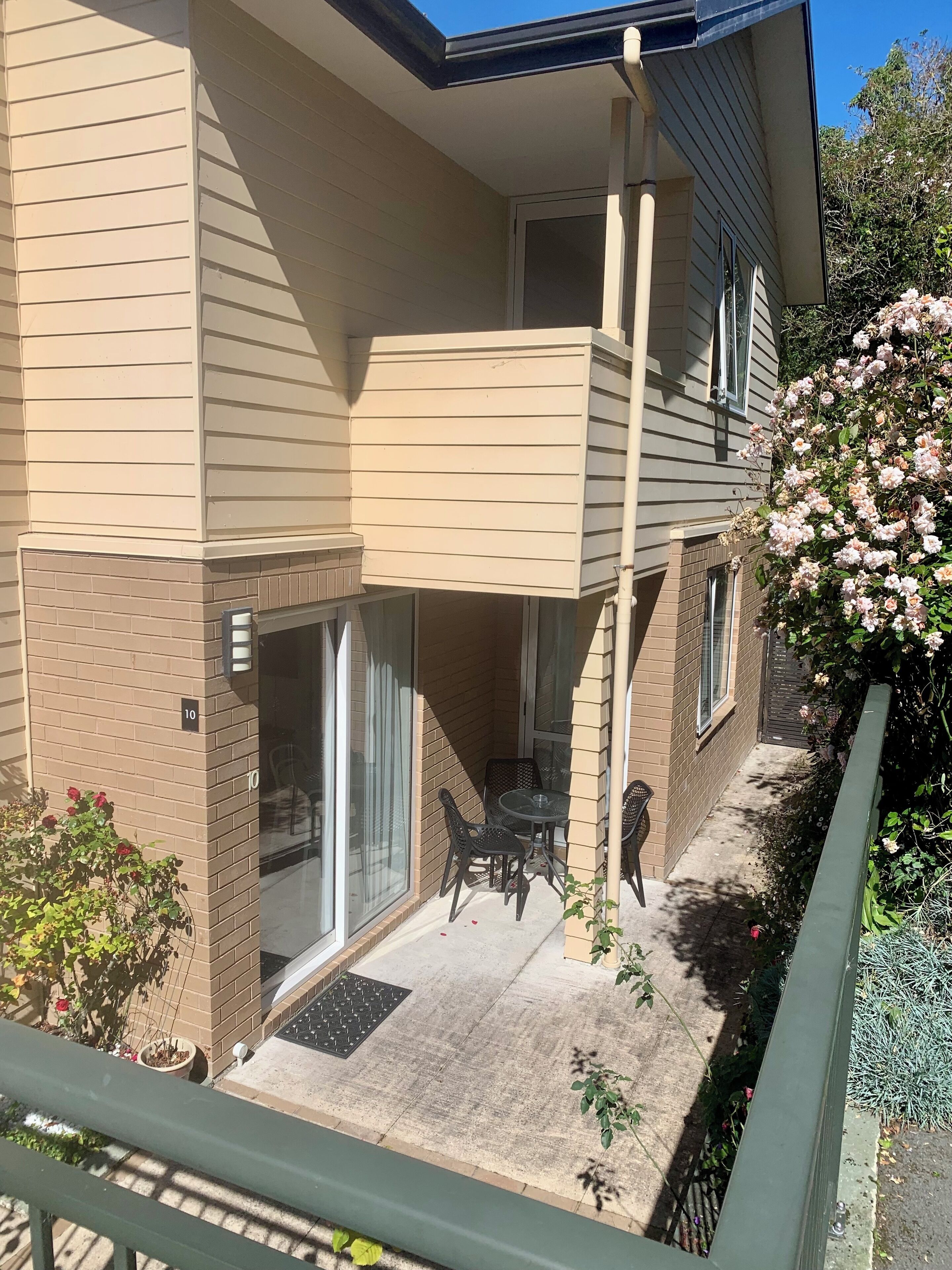 Un bâtiment à deux étages doté d’un balcon, d’une porte donnant sur un patio couvert et d’un petit jardin agrémenté de fleurs.