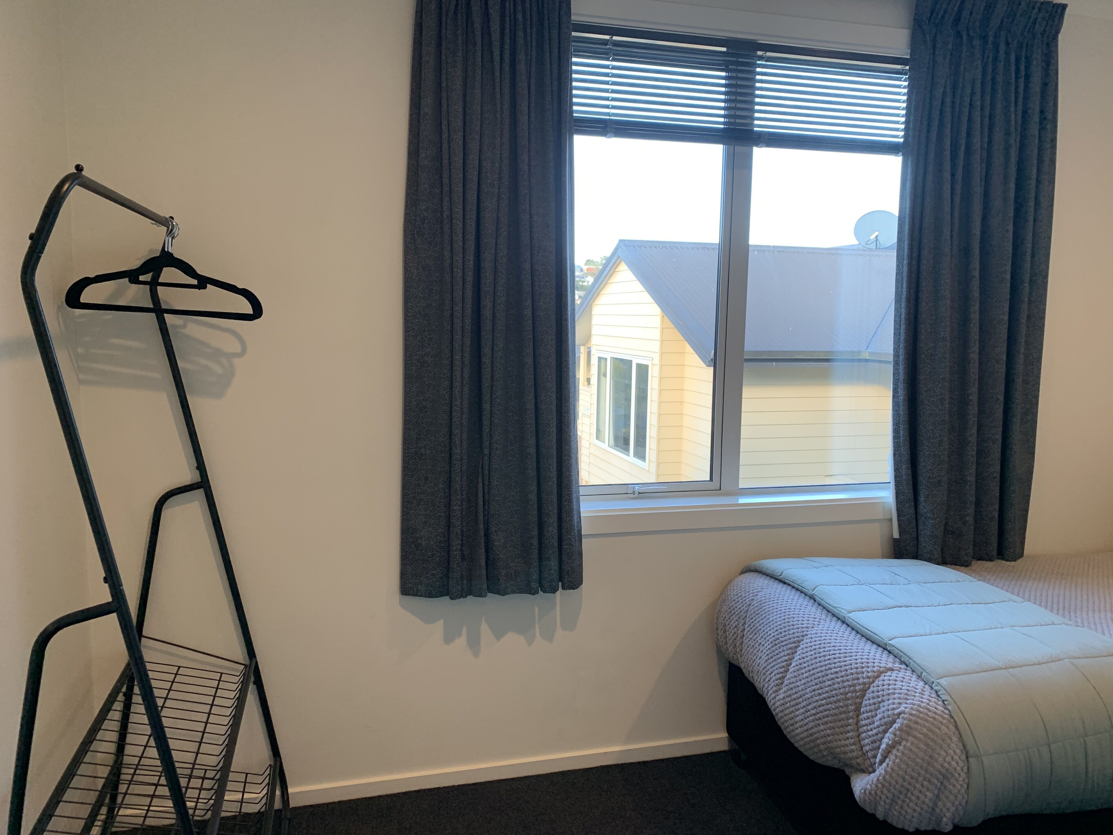 Un dormitorio con una cama individual, una ventana con cortinas y un tendedero contra la pared.