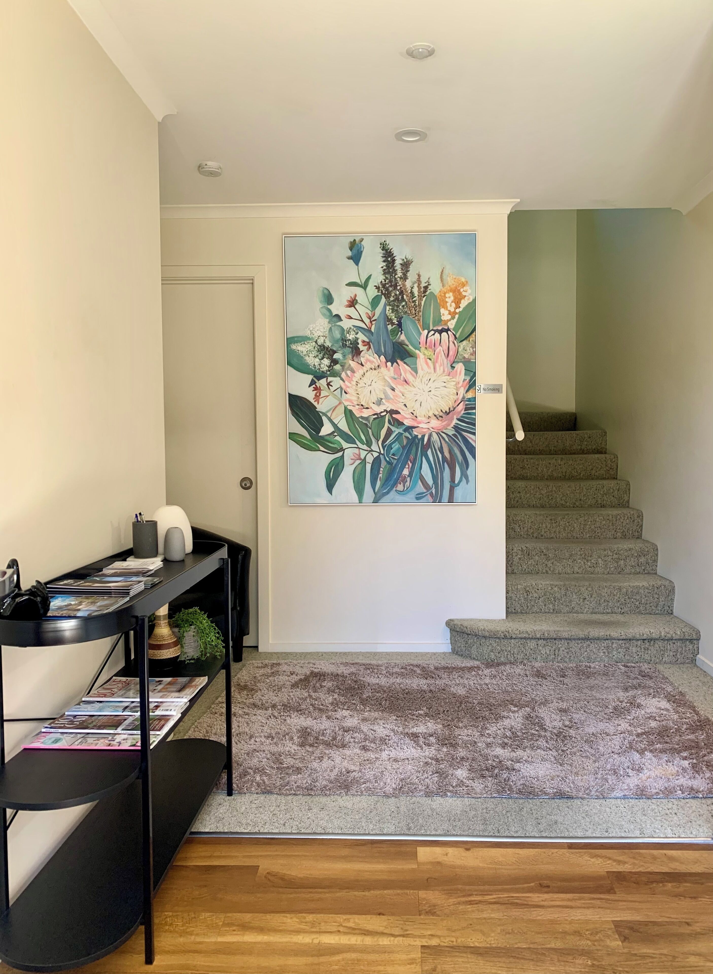 A modern interior with a wooden floor, a black console table with magazines, a large floral painting, and a staircase.