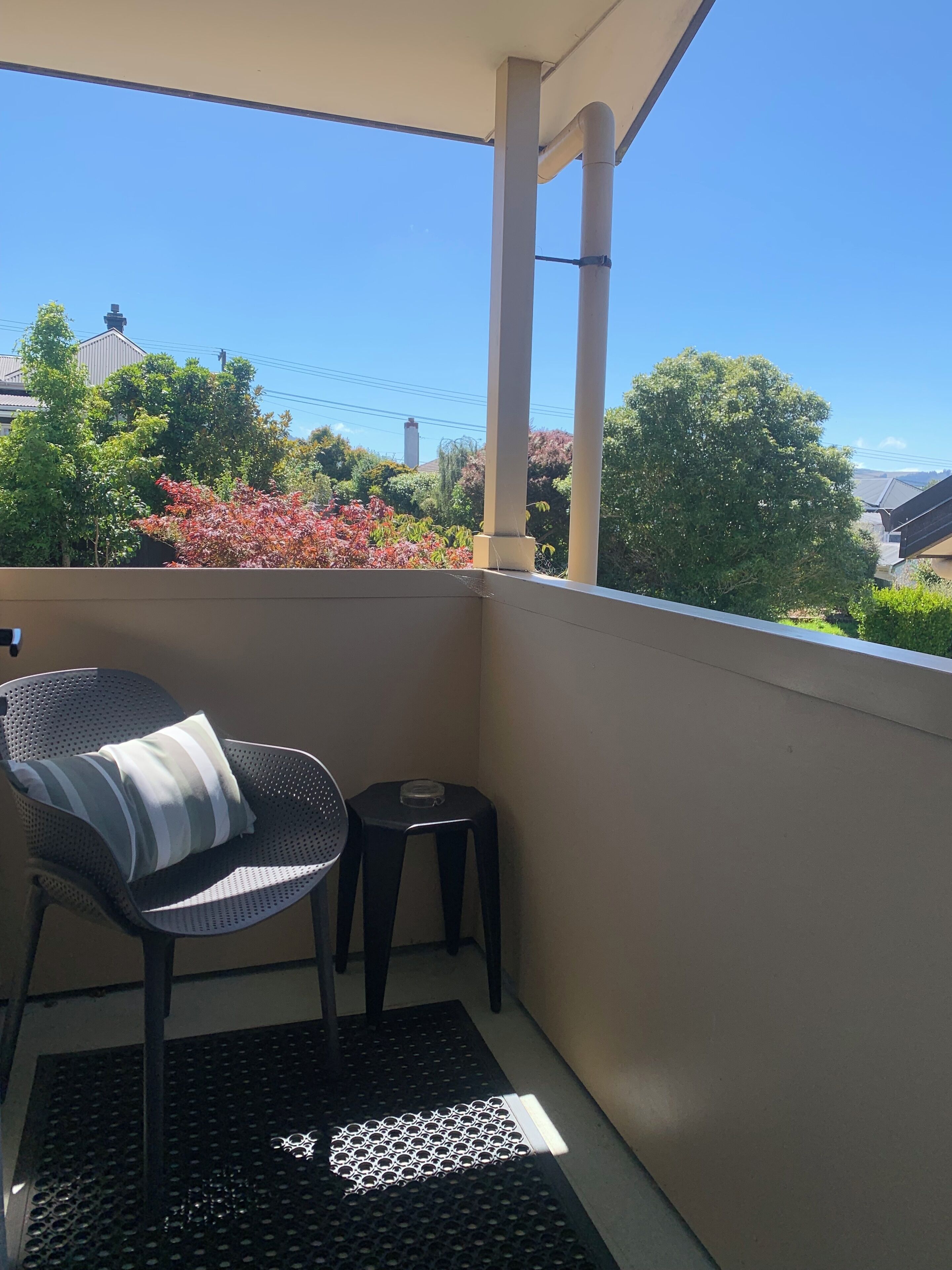 A balcony with a chair and a small stool, a metal grate floor, and a view of trees and buildings.