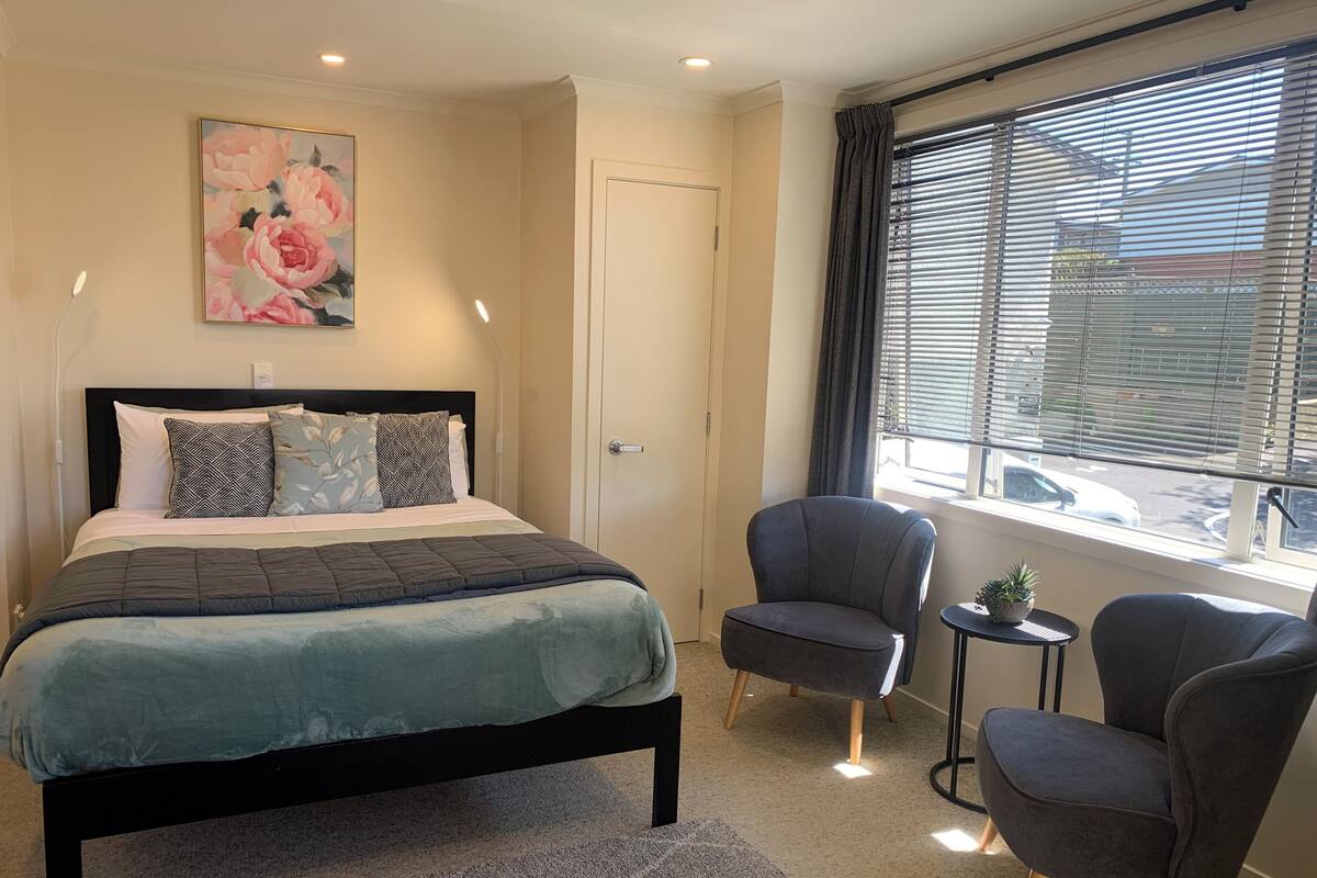 A bedroom with a bed, two armchairs, a small table, a painting, and a window with blinds.