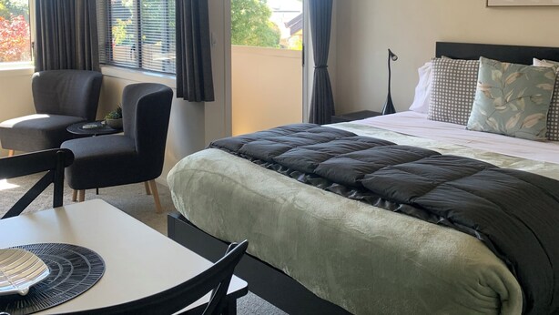 A hotel room with a bed, a desk, and two chairs. There is a window with curtains, and a plant on the desk.