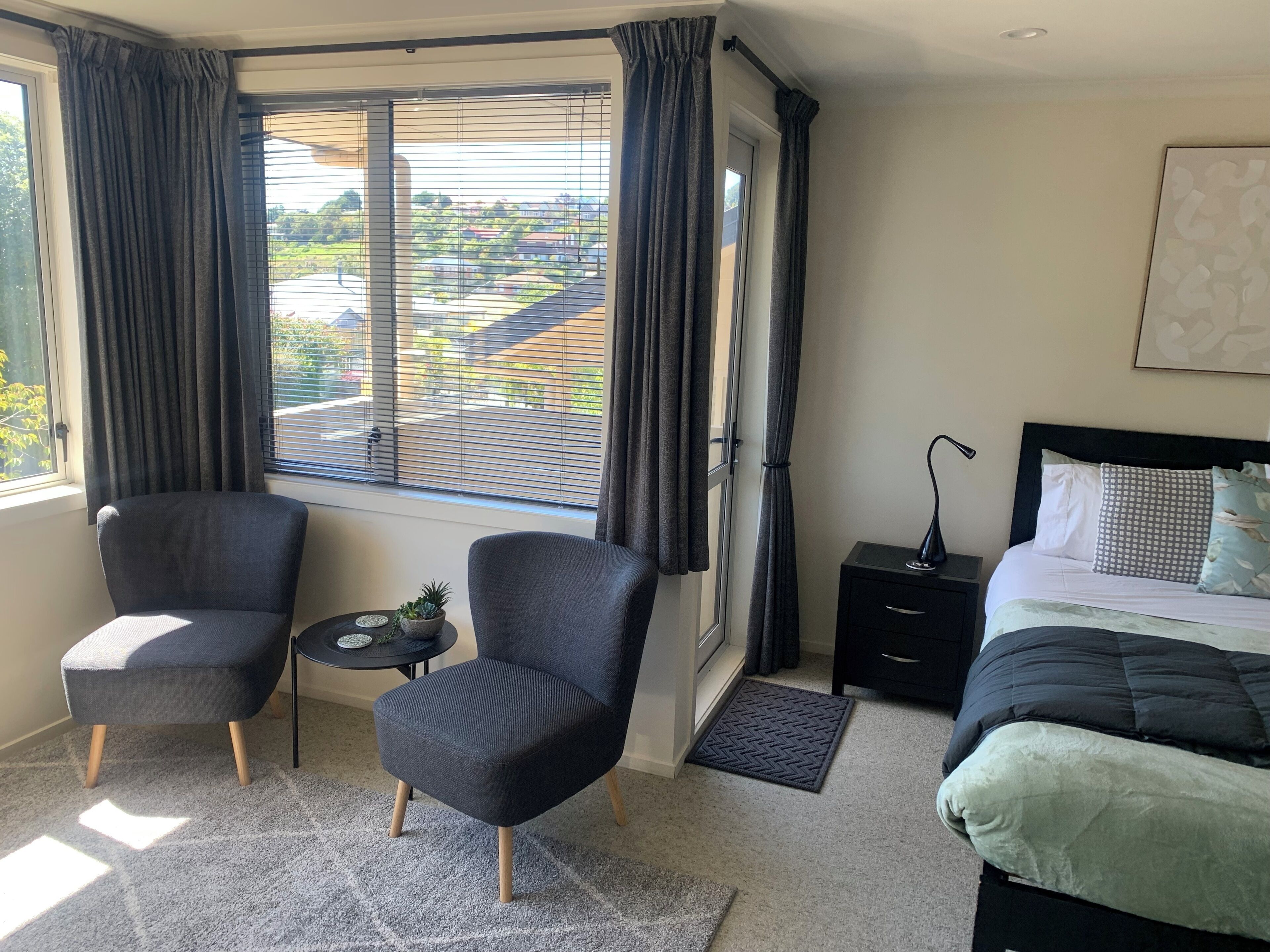 Un dormitorio con dos sillas, una mesita de noche, una lámpara, una cama y vistas a una zona residencial a través de amplias ventanas.