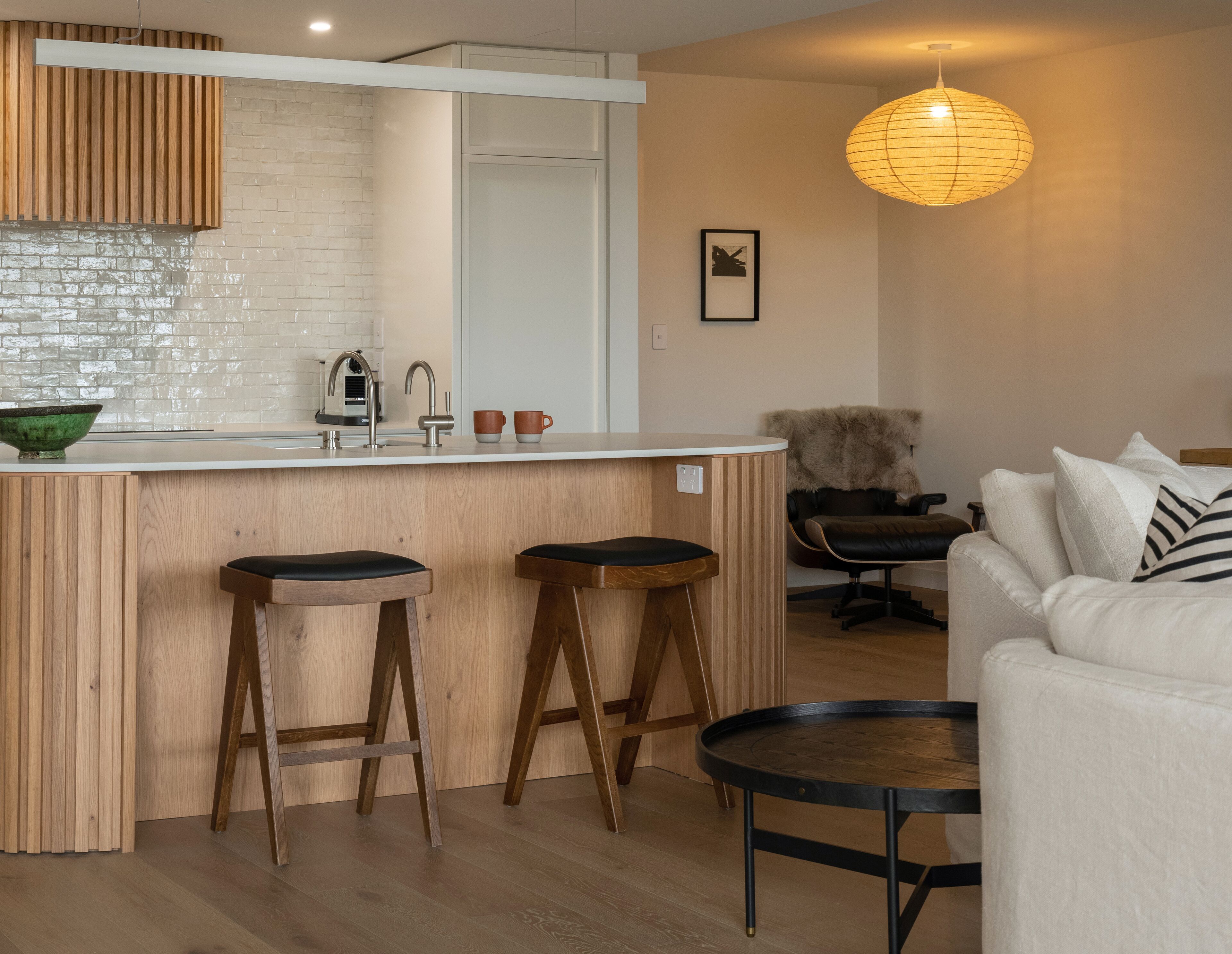 A modern kitchen with a white countertop, wooden stools, a hanging light fixture, and a white sofa in a living area.