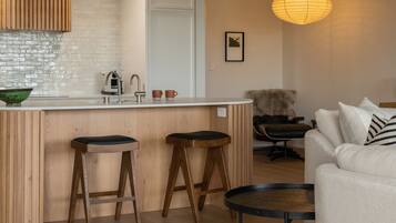 A modern kitchen with a white countertop, wooden stools, a hanging light fixture, and a white sofa in a living area.