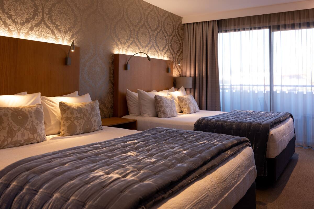 A hotel room with two beds, a large window with curtains, and a patterned wallpaper.