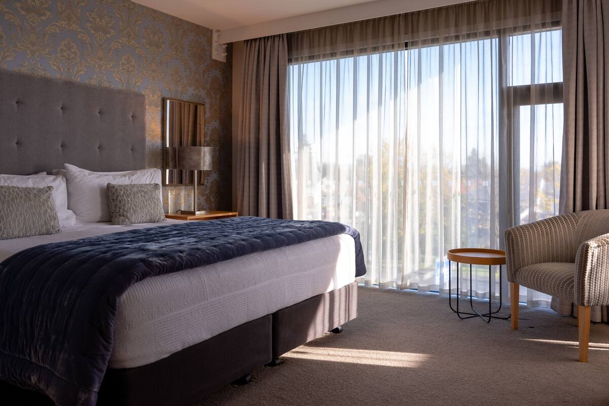 A hotel room with a large bed, a chair, a nightstand, and a window with curtains.