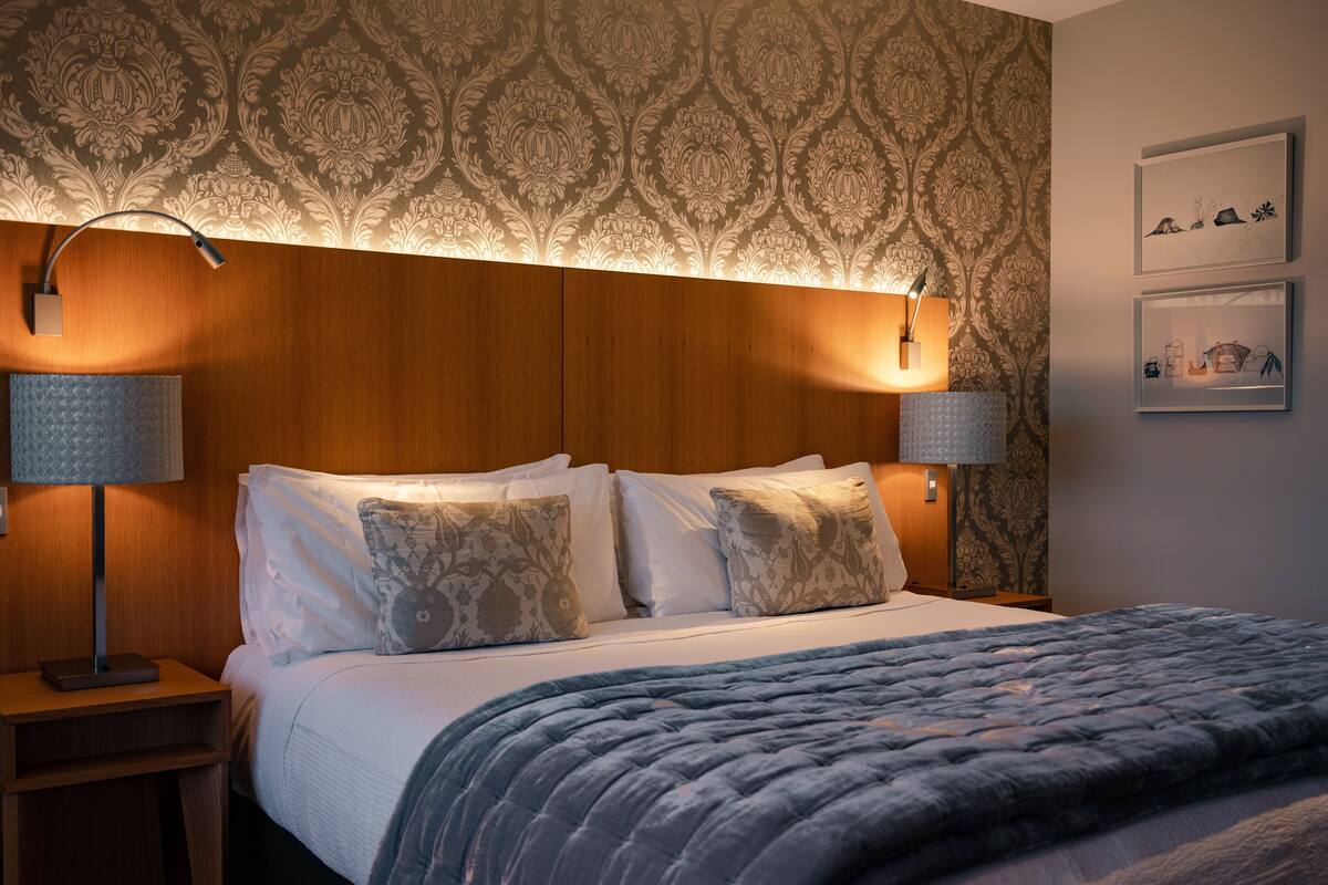 A neatly made bed with a patterned blanket, two bedside lamps, and a wall with a decorative wallpaper pattern.
