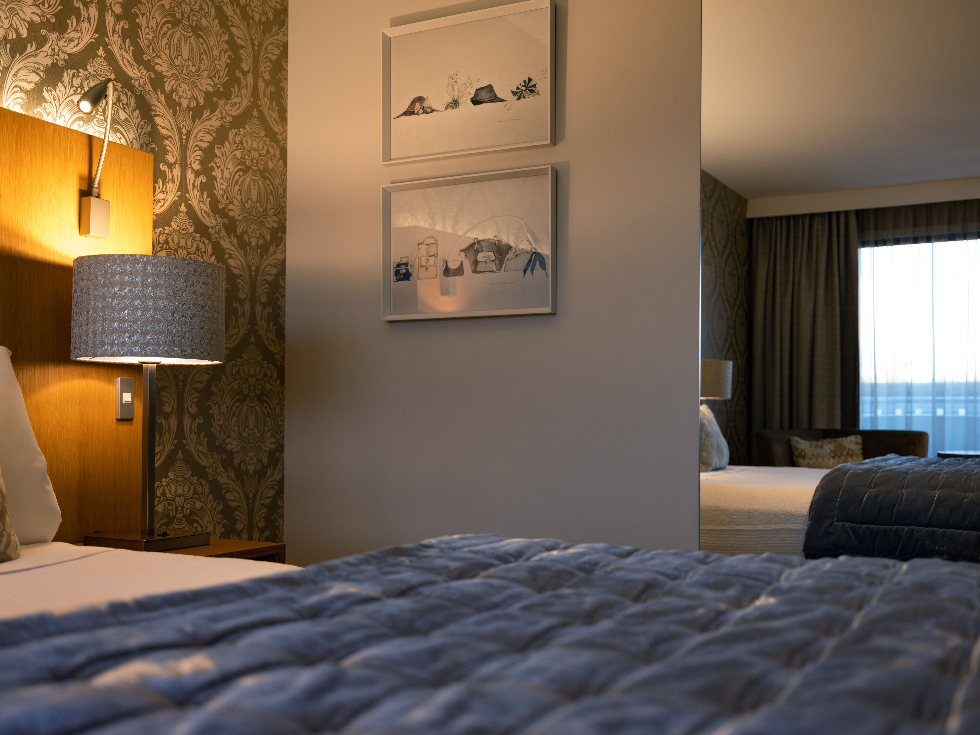 A hotel room with a bed, a desk, a lamp, and two framed pictures on the wall.