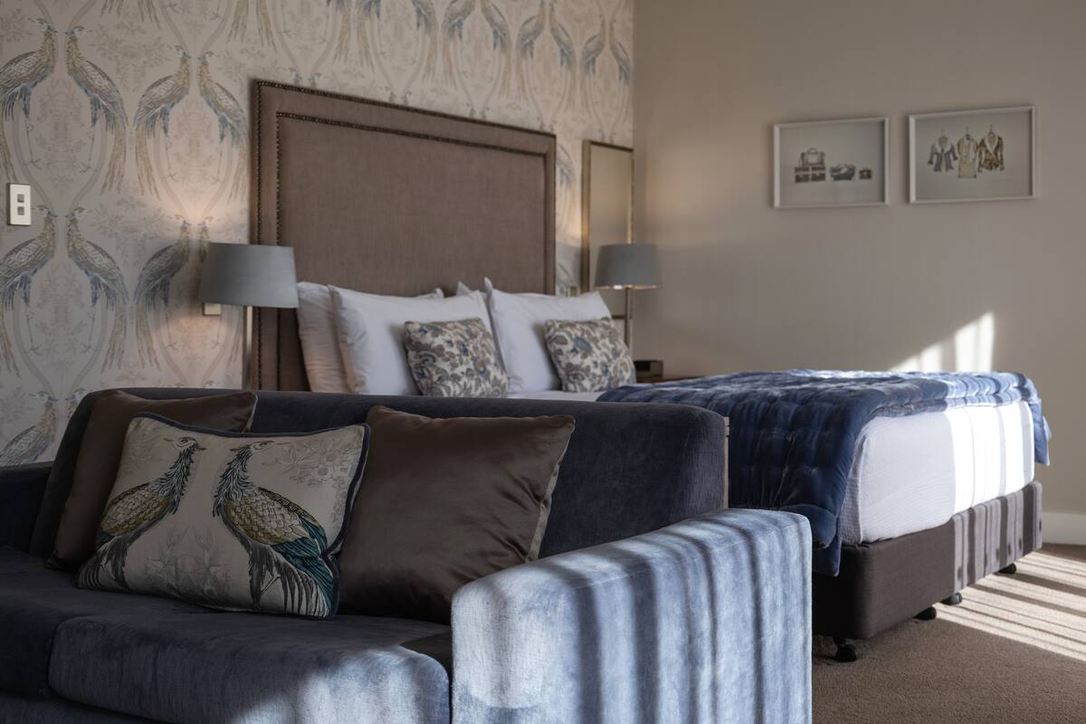 A hotel room with a large bed, a sofa, and a patterned wallpaper. There are two bedside lamps and a framed picture on the wall.