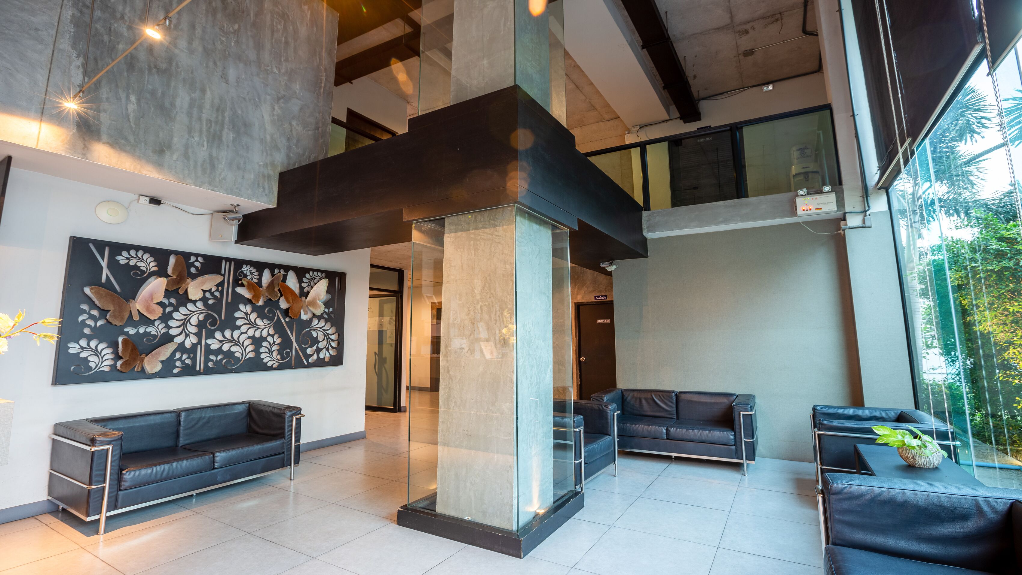 A modern lobby with a glass partition, a black sofa, a decorative butterfly wall art, and a view of greenery outside.