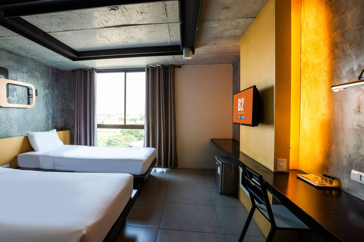 A hotel room with two beds, a desk, a TV, and a window with curtains.