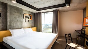 A hotel room with a large bed, a desk, a television, and a view of the city through the window.