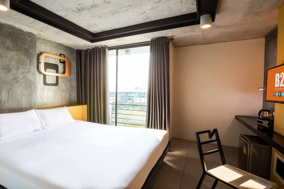 A hotel room with a large bed, a desk, a television, and a view of the city through the window.