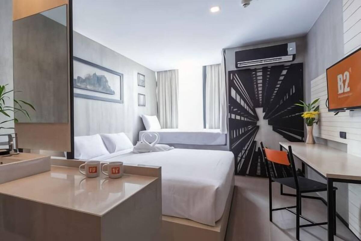 A modern hotel room with a bed, a desk, a chair, a mirror, a wall-mounted TV, and a window with curtains.