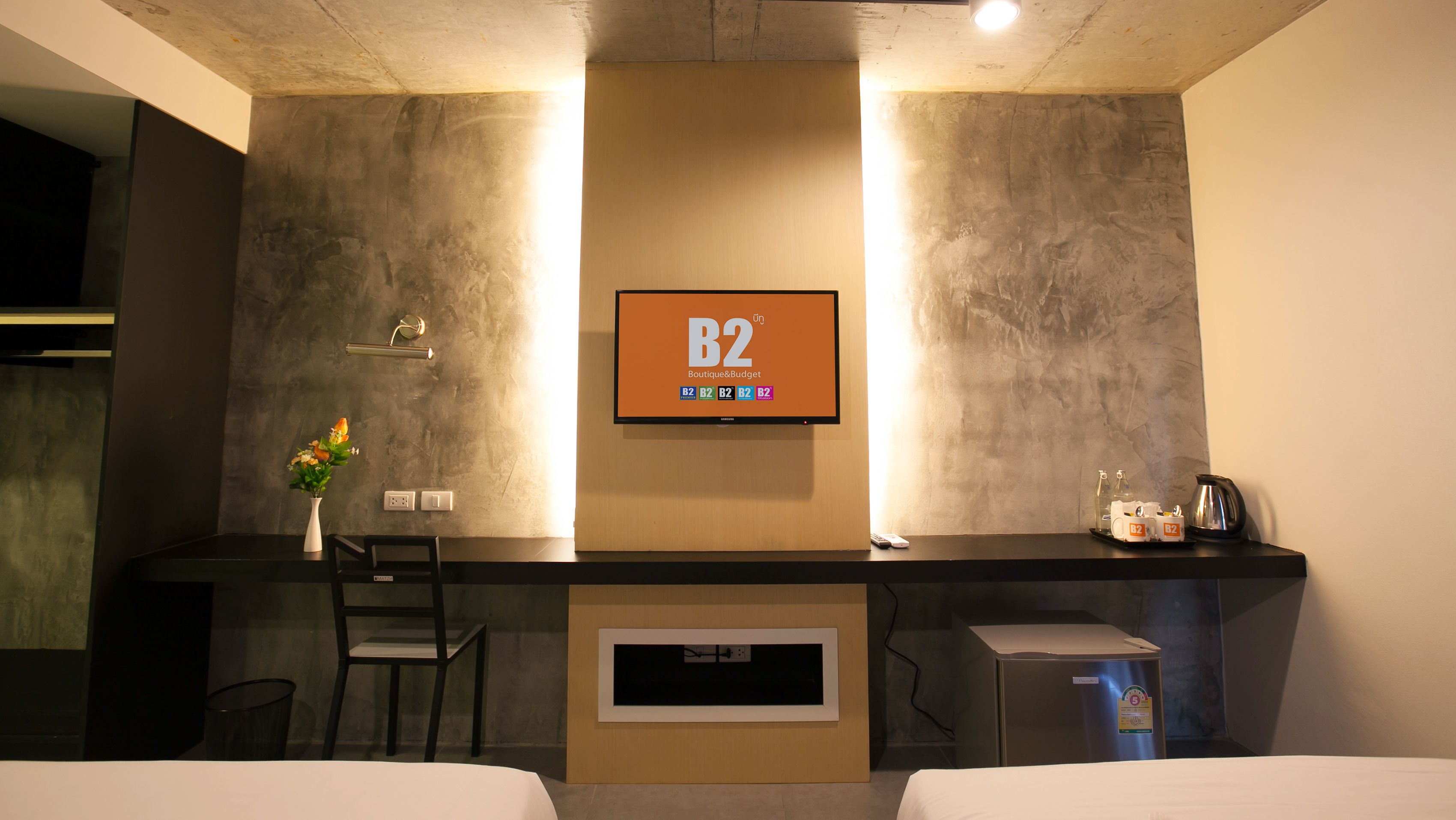 A hotel room with a bed, a desk, a chair, a TV mounted on the wall, and a small refrigerator.